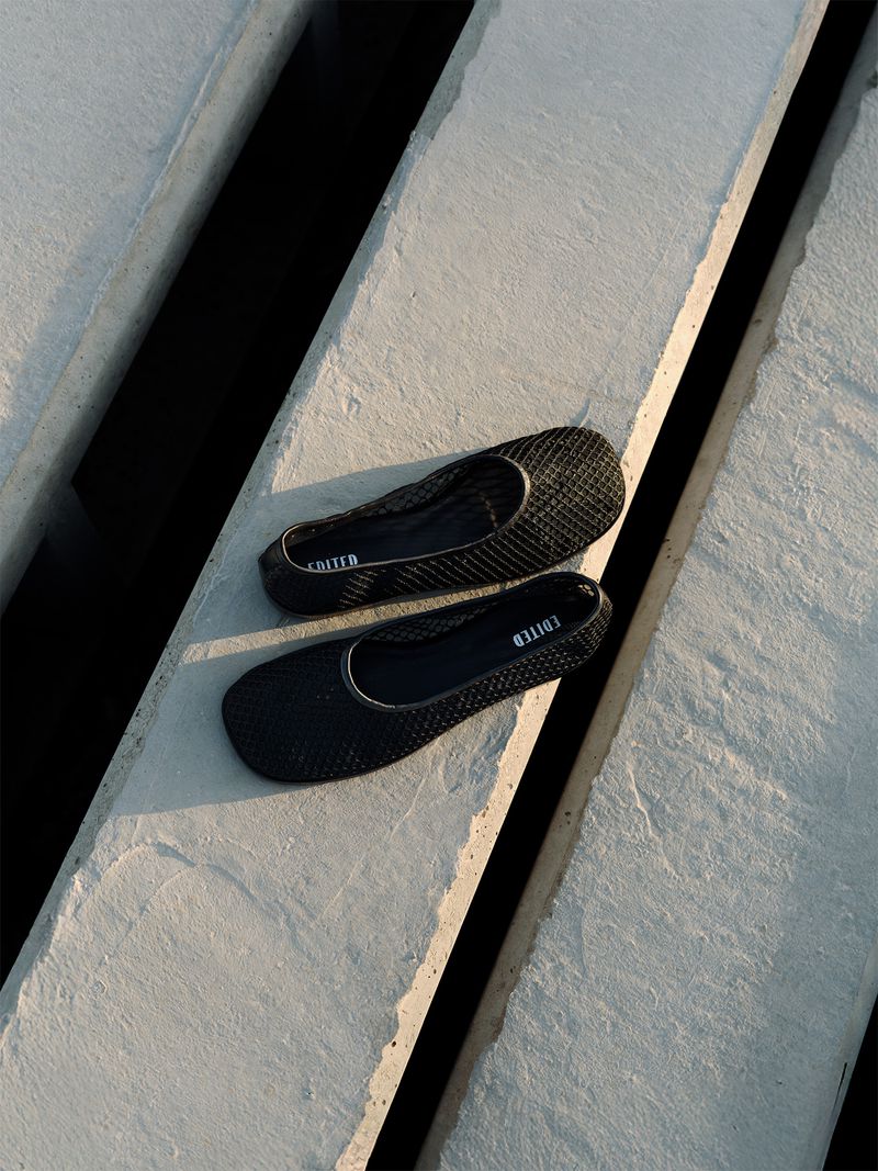 A pair of black ballet flats are on a concrete surface with linear shadows, capturing a minimalist fashion aesthetic with a soft, warm tone.