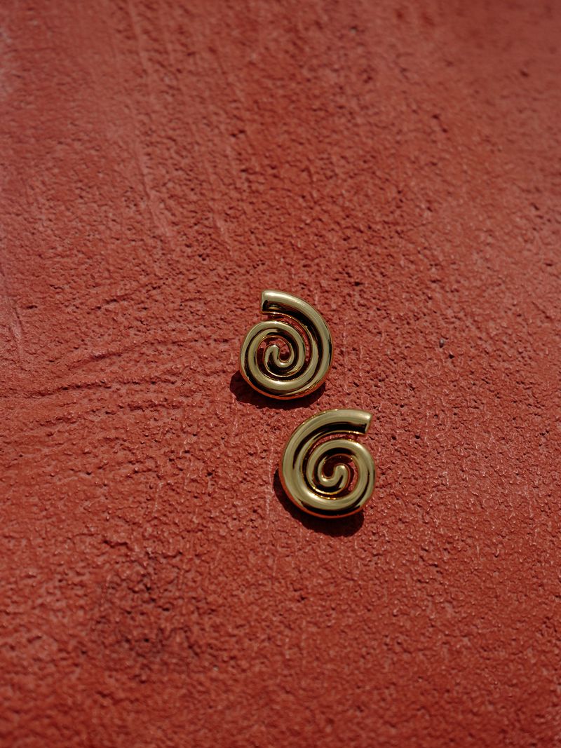 Close-up of gold spiral earrings on textured red background showcasing minimalist jewelry design and warm color palette in a studio setting.