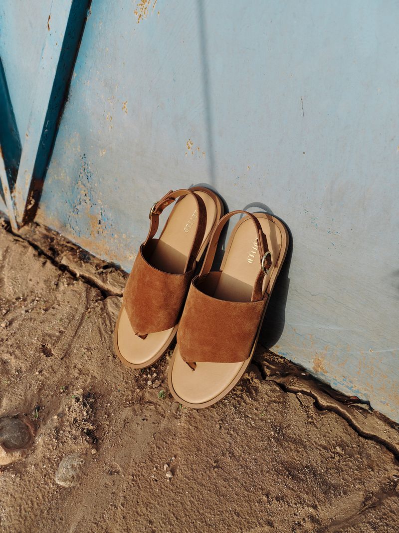 Suede sandals with beige soles sit on concrete near a blue wall, showcasing a fashion-forward style with earthy tones and textured surfaces.