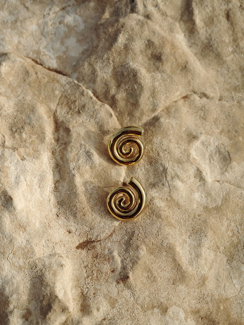 Close-up of two gold-colored spiral earrings resting on a textured beige stone surface, illuminated by natural light.