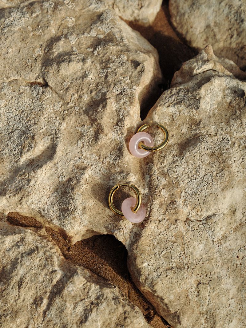 Close-up shot of pink stone and gold hoop earrings on a textured rock surface, showcasing minimalist jewelry design.