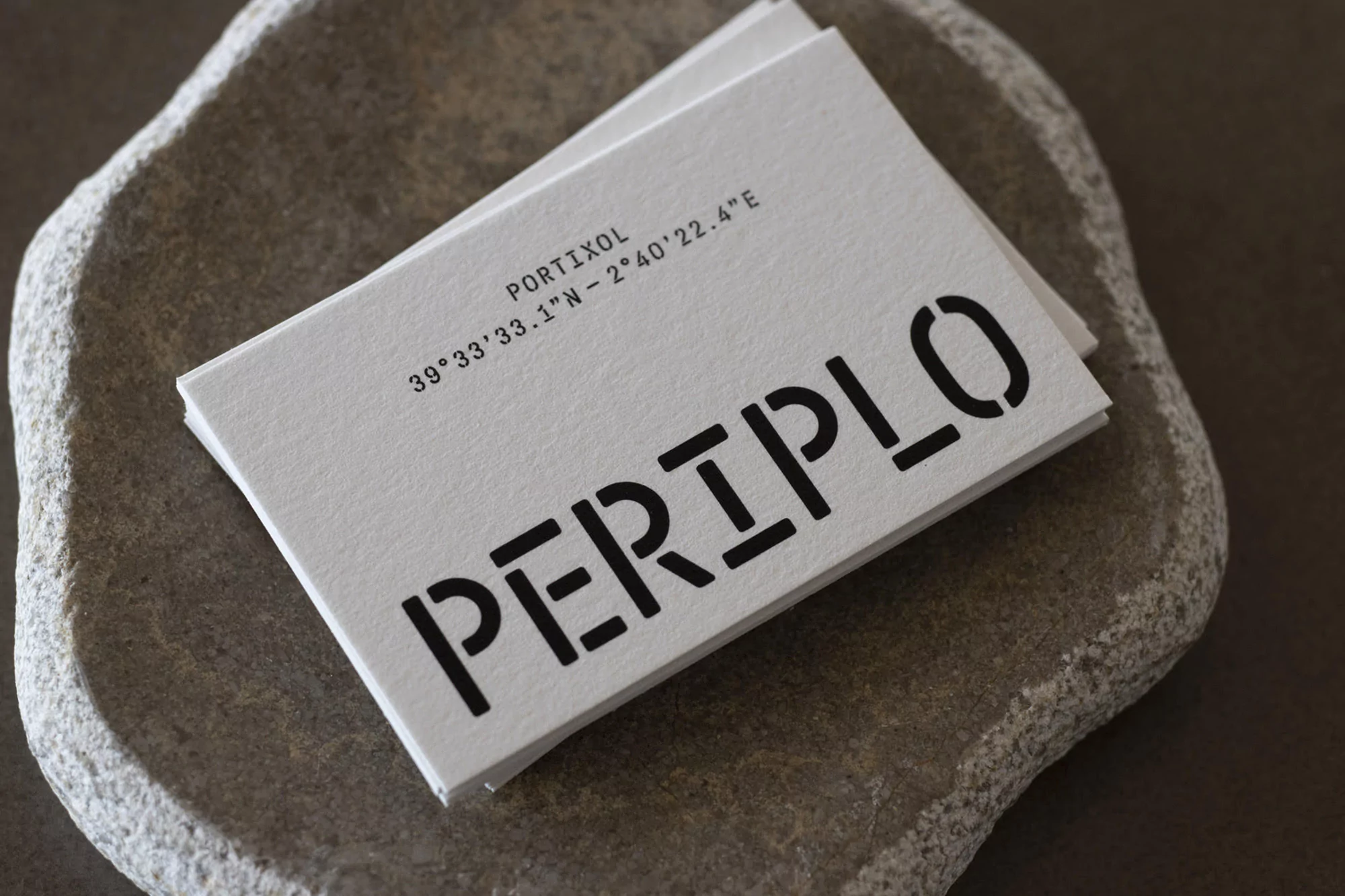 Close-up of stacked business cards on a textured stone surface, featuring stencil-style typography and geographic coordinates in black ink.