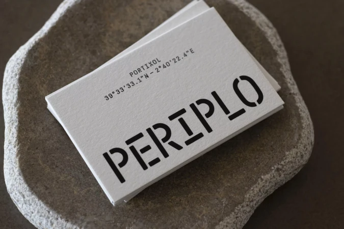 Close-up of stacked business cards on a textured stone surface, featuring stencil-style typography and geographic coordinates in black ink.