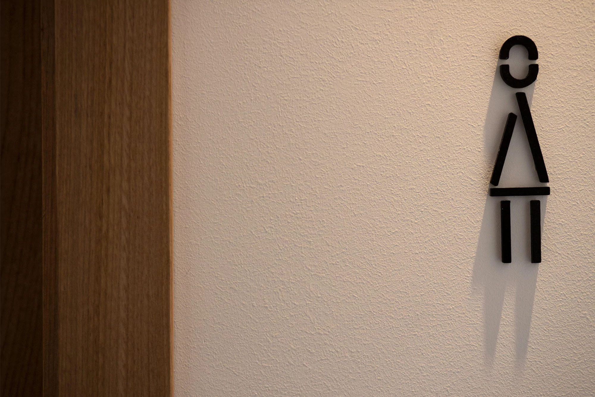 Restroom sign on a beige wall next to a dark wooden door. The sign is a stylized woman figure, indicating a female restroom.