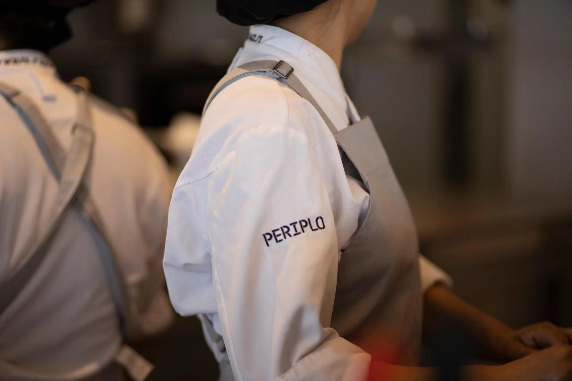 Two chefs stand side-by-side in a kitchen wearing white coats and grey aprons. The word "PERIPLO" is stitched on one coat sleeve.