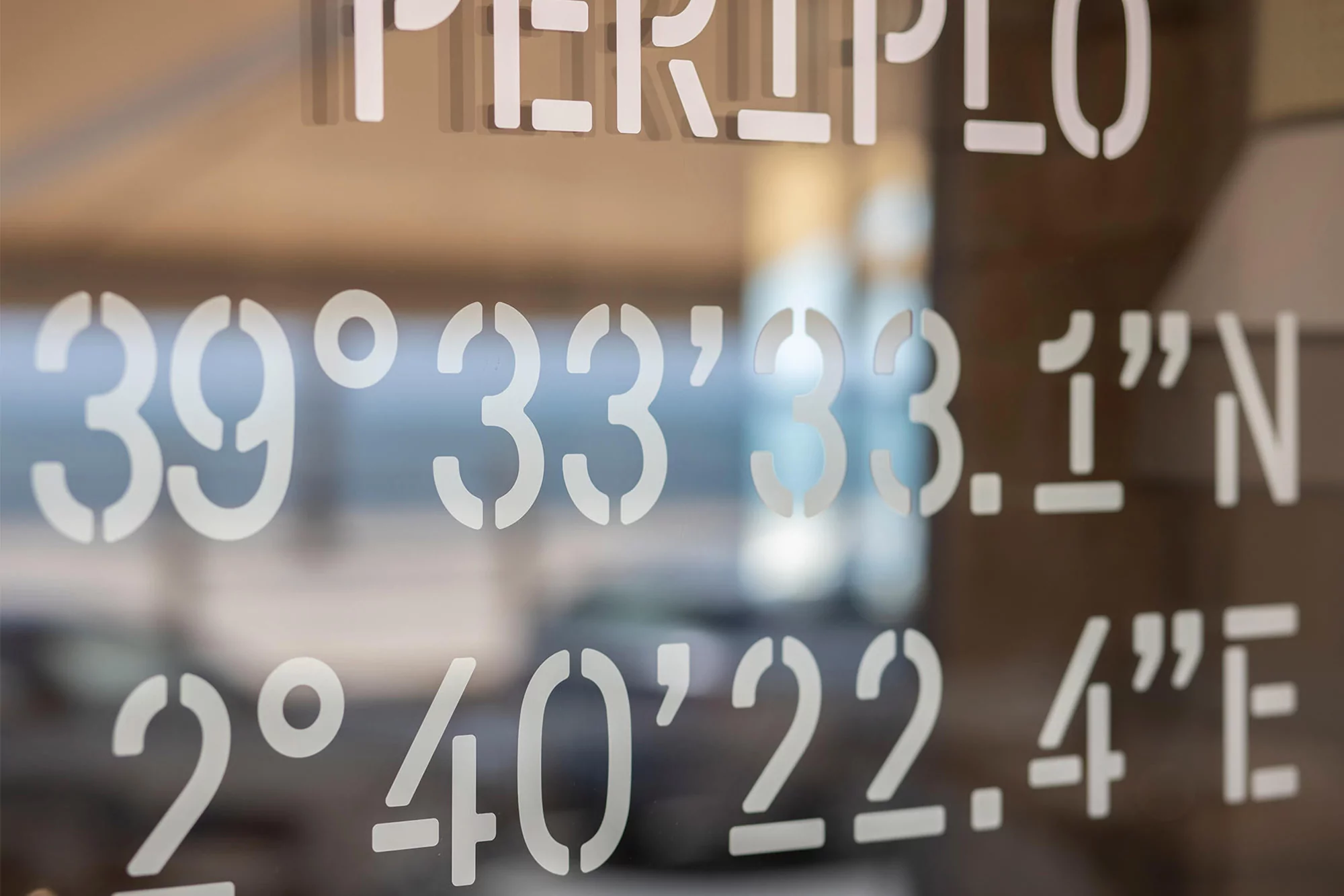 White text on glass shows coordinates 39°33'33.1"N, 2°40'22.4"E, with blurred background. Lettering is PERIPLO at the top.