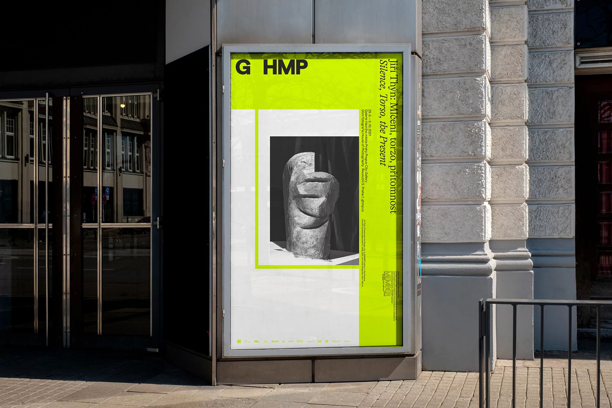 Outdoor advertisement features a head sculpture on a poster with bright yellow accents, displayed next to a modern building entrance.