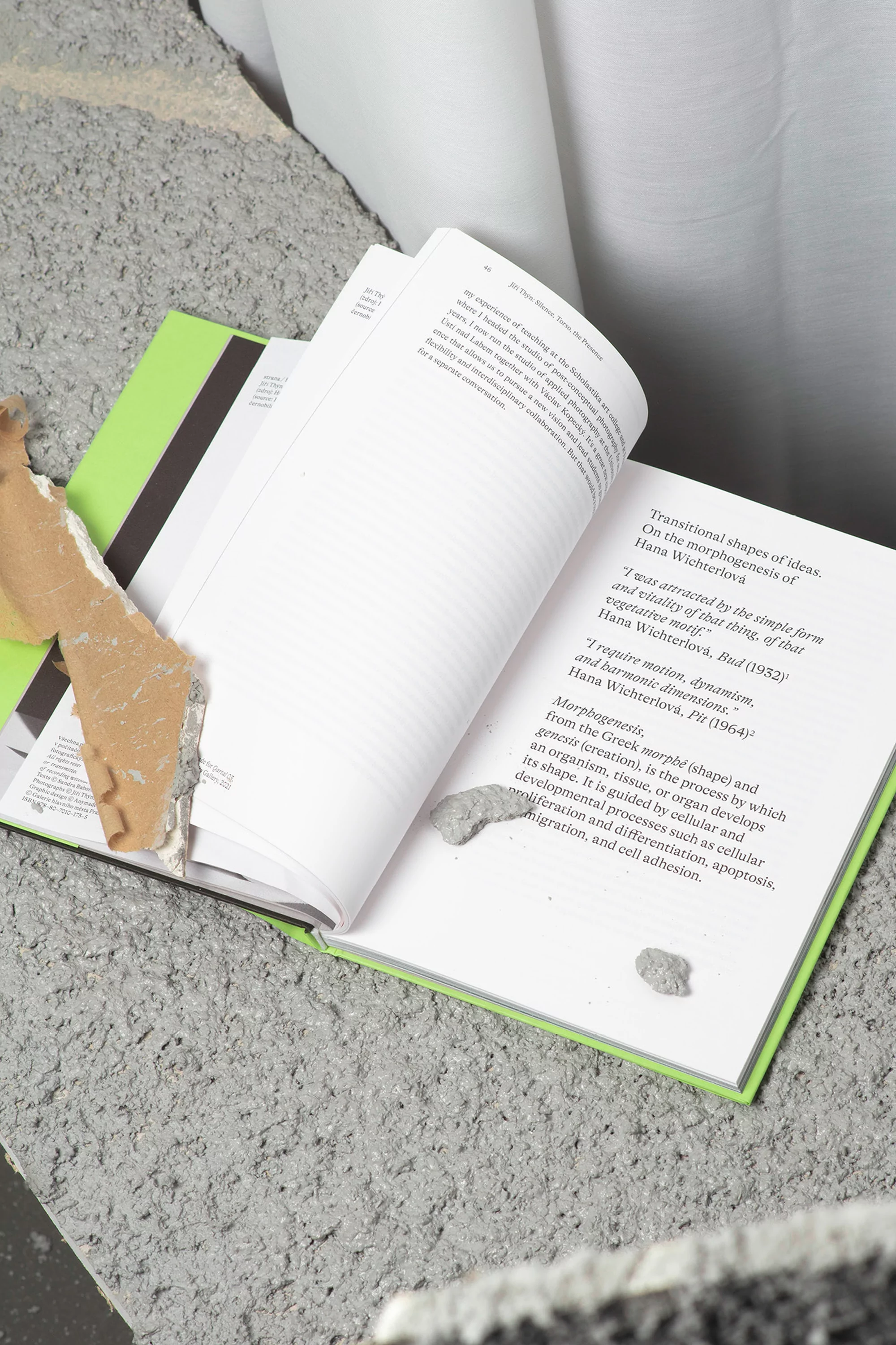 Open book with text and a neon green cover rests on textured gray material, alongside a white sheet and torn cardboard pieces.