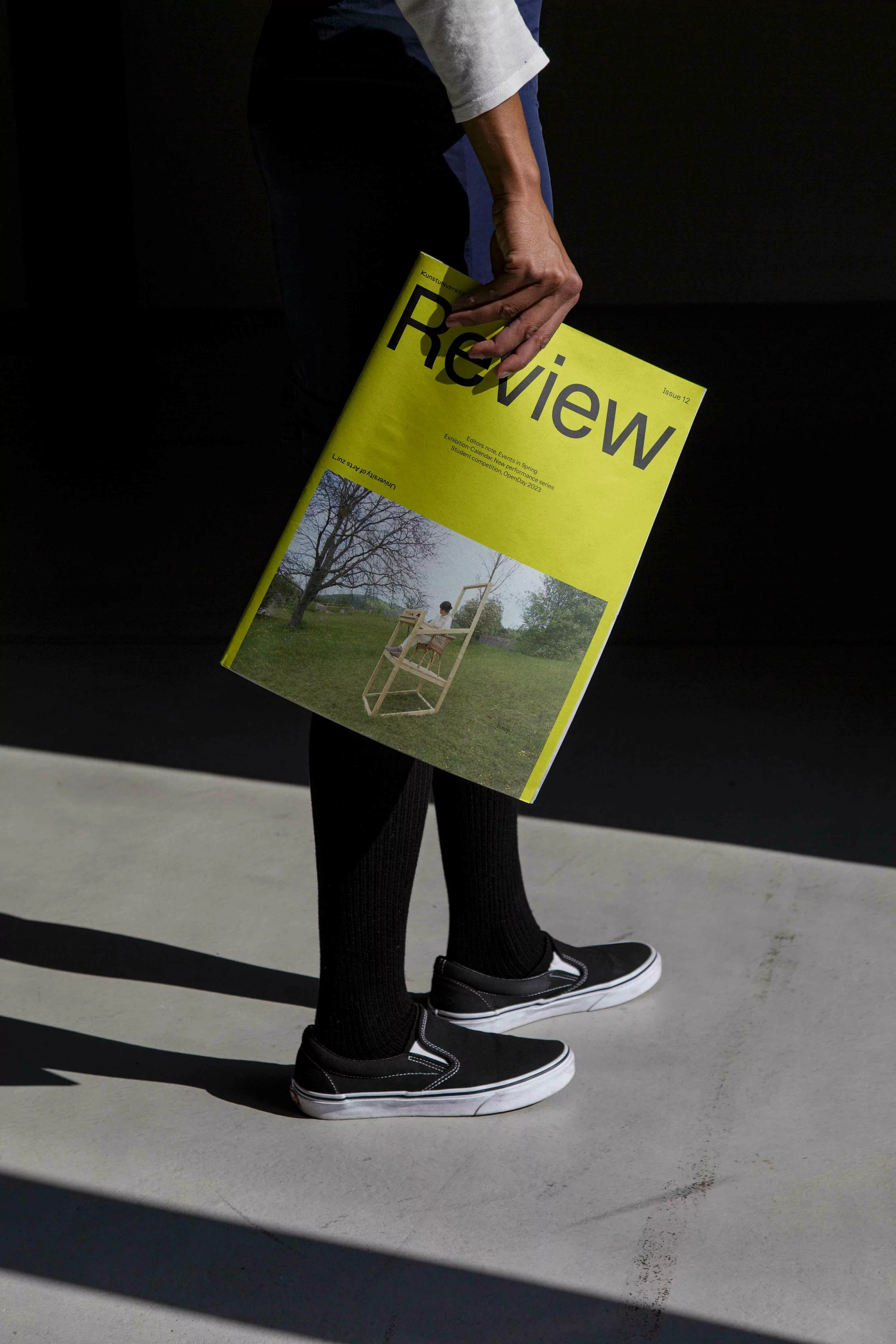 Person holding a yellow Review magazine with a lawn chair cover photo, paired with black-and-white slip-on shoes on a grey surface.