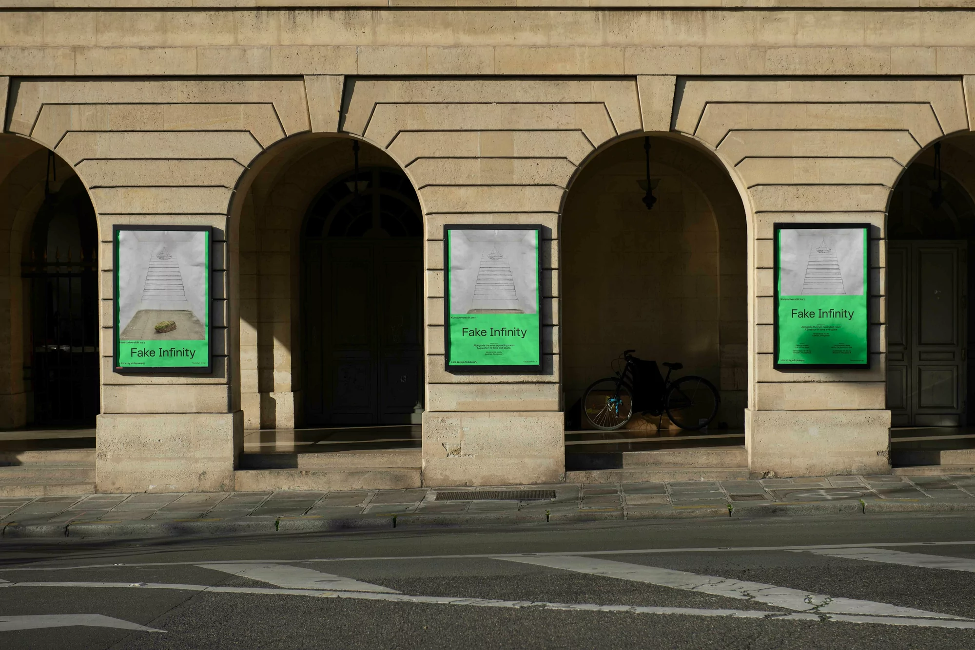 Three posters with green trim line an arched building facade with a bicycle parked in the background on a city street in daylight.