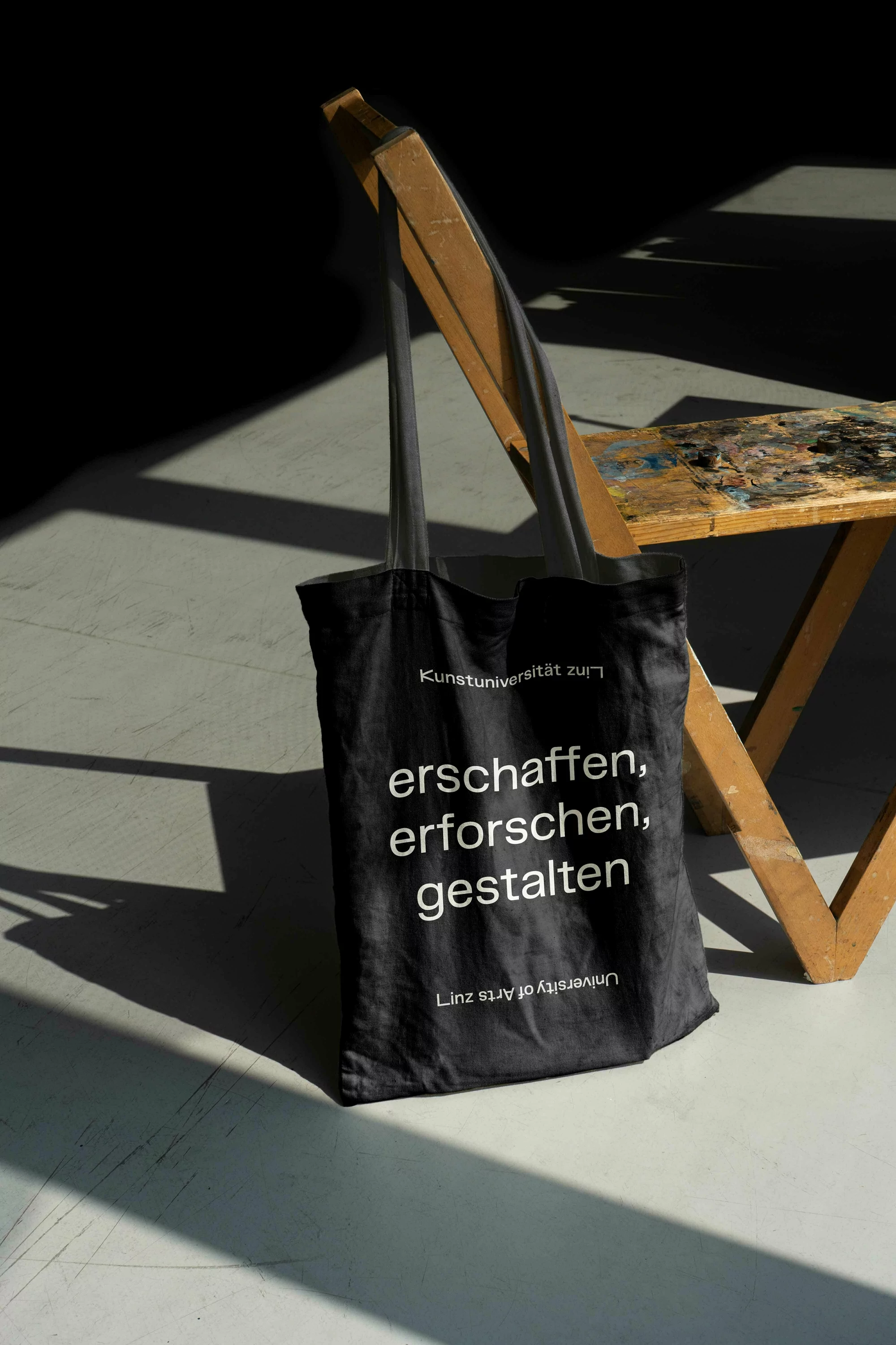 Black tote bag hanging on a wooden chair in a brightly lit room. The bag has white text printed on it. 