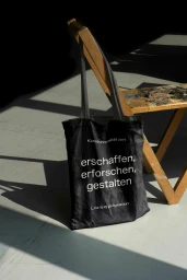 A black canvas tote bag with white typography hangs on a wooden chair, casting long diagonal shadows across a light floor.