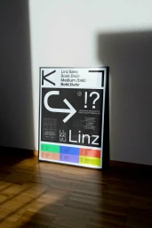 Framed typographic poster leaning against a plain wall on a wooden floor, illuminated by angled natural light casting shadows.