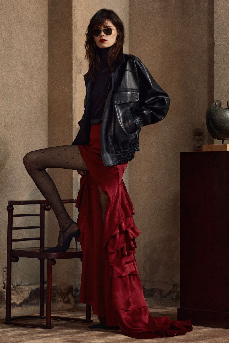 Elegant woman in a black leather jacket, red ruffle dress, and polka dot stockings sits on a wooden chair in a studio setting.