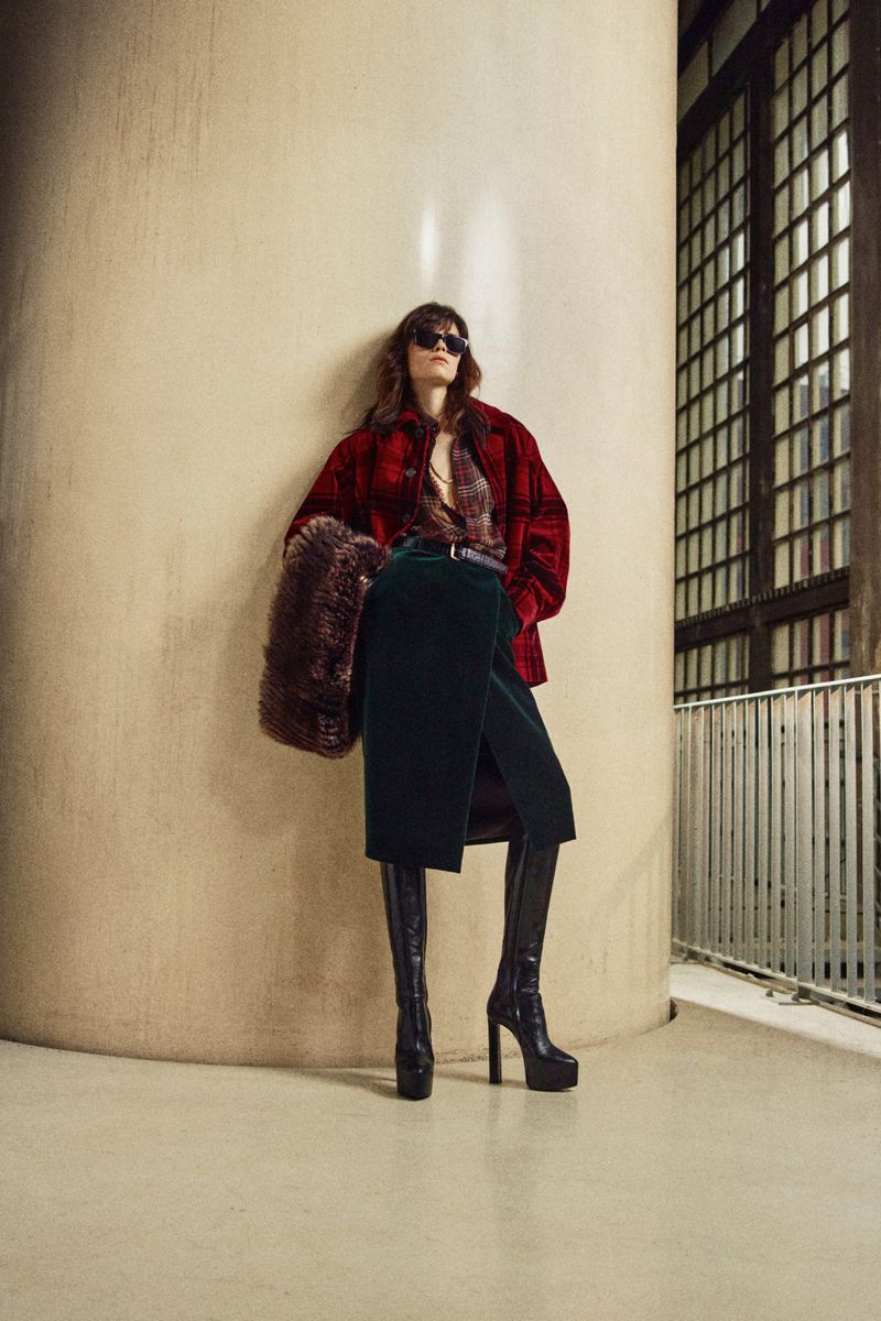 A stylish woman with long brown hair in a red jacket and dark green skirt poses in front of a cream-colored pillar and iron gate.
