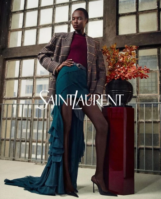 A Black female model showcases Saint Laurent fashion in an indoor setting with a window backdrop and a vase of flowers.