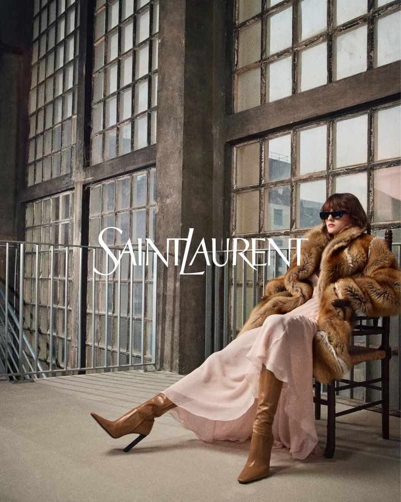 A woman in a fur coat poses in an industrial-style interior next to large windows for a Saint Laurent fashion advertisement.