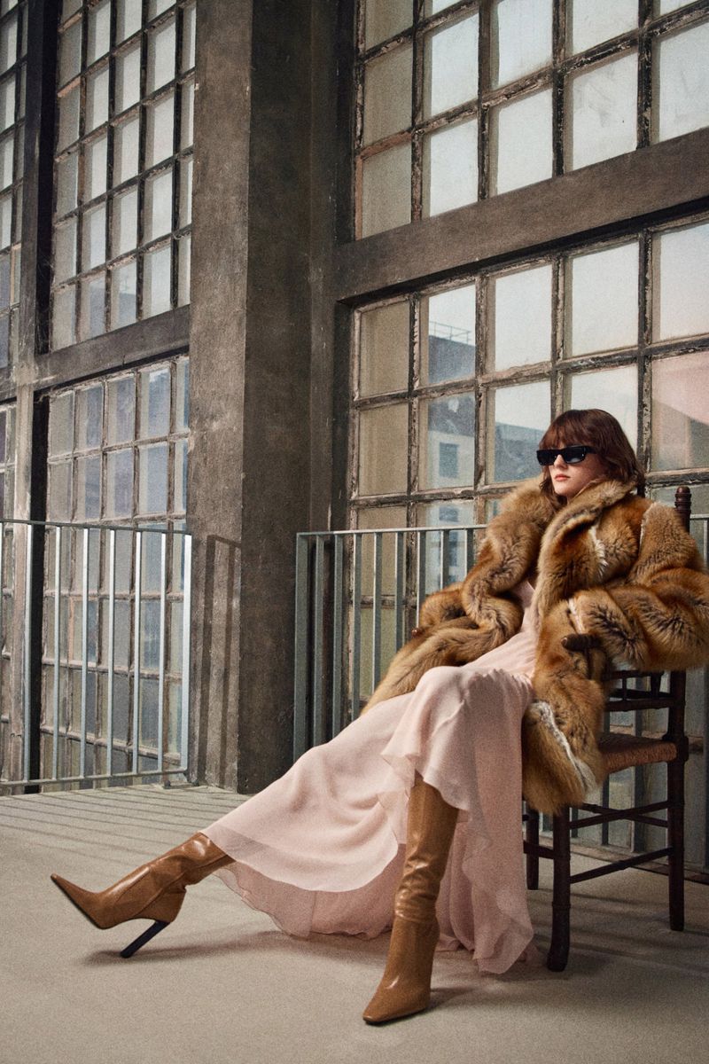 A woman in a fur coat, dress, and boots sits on a chair indoors near large windows, with diffused lighting casting soft shadows.