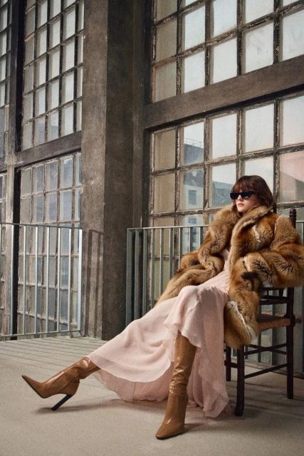 A woman in a fur coat, dress, and boots sits on a chair indoors near large windows, with diffused lighting casting soft shadows.