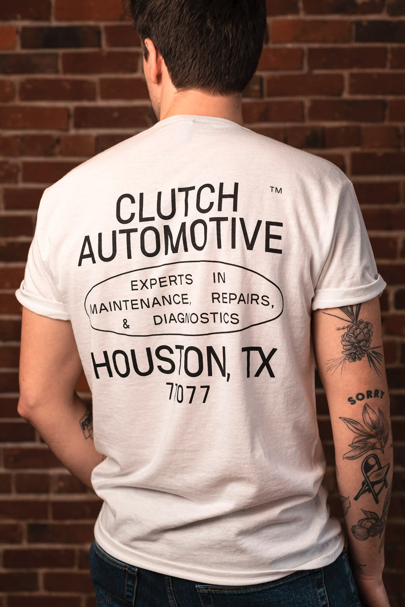 Man wears a Clutch Automotive T-shirt in front of a brick wall, showing automotive maintenance repair logo and Houston, TX text.