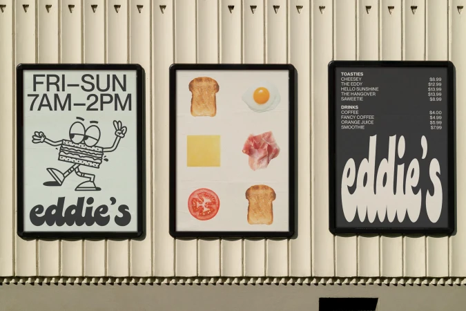 Three framed posters hang on a wall, showcasing restaurant information, menu item illustrations, and toastie/drink options with prices.