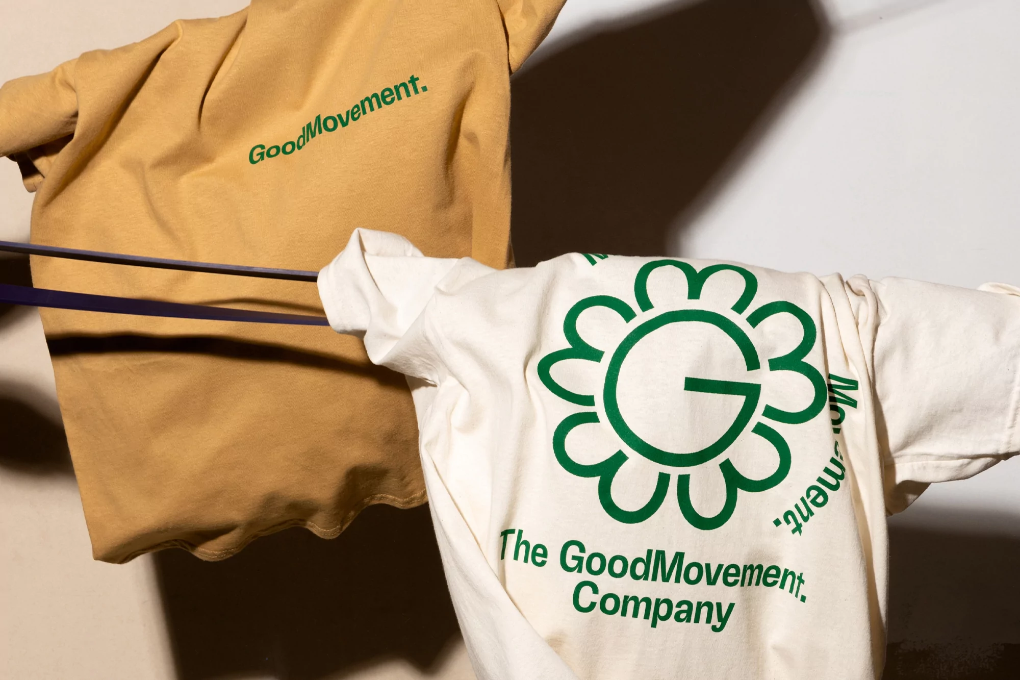 Two GoodMovement branded T-shirts, one beige and one white, hang against a wall, showcasing the logo designs in a product shot.