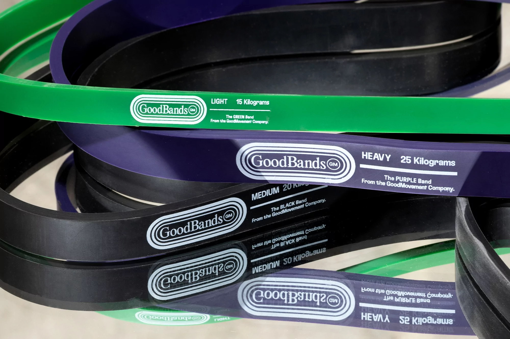 Close-up of layered resistance bands in green, purple, black, and grey with branding and weight indicators. Fitness and exercise equipment.