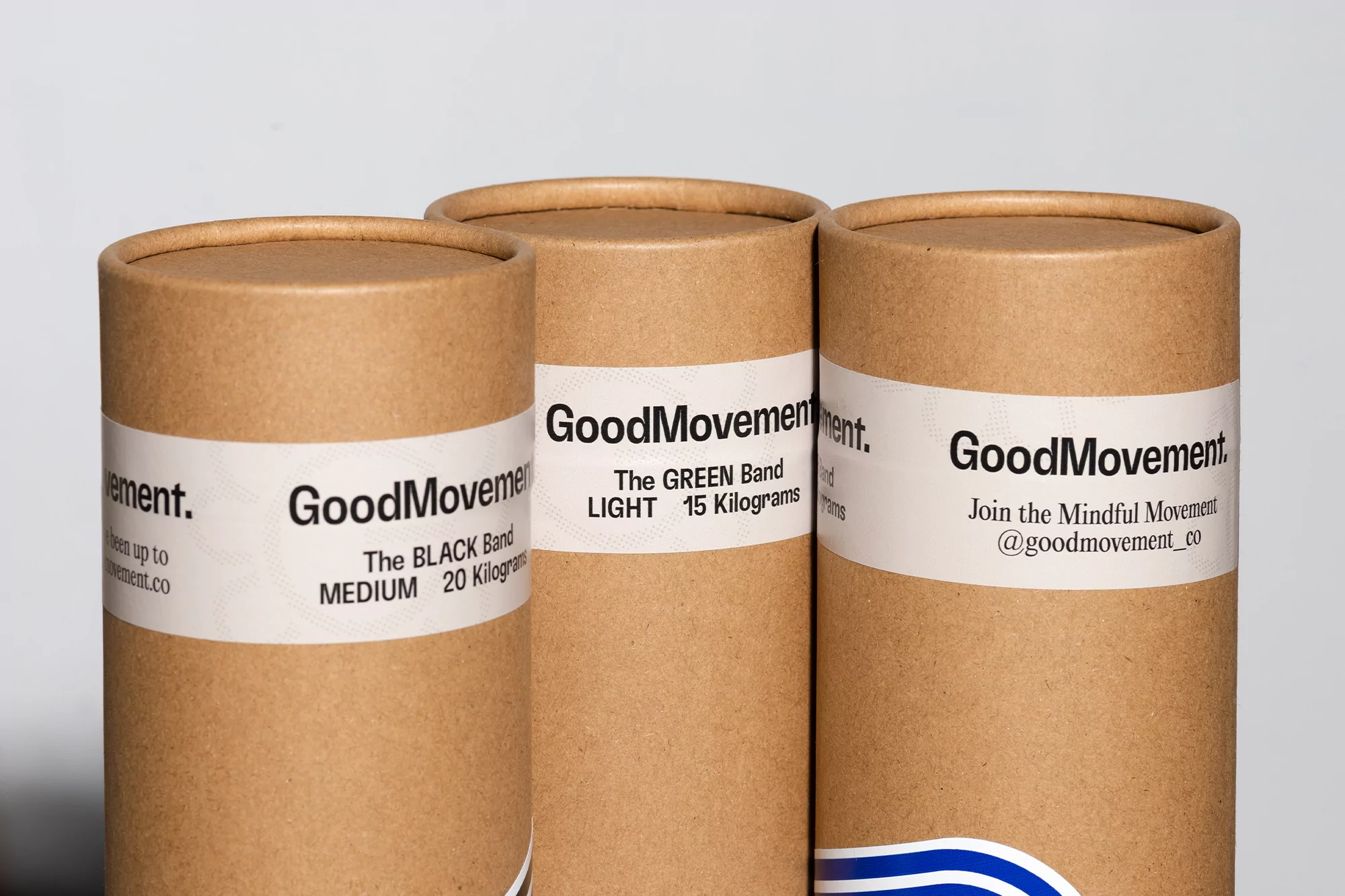 Three cardboard tubes of GoodMovement resistance bands displaying band level and weight on white labels, indoor setting.