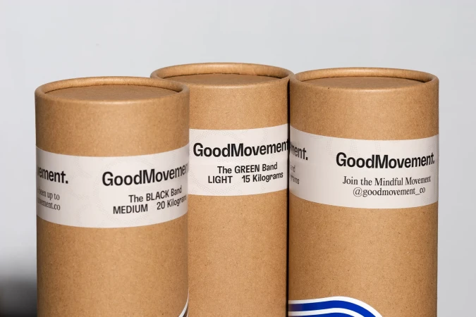 Three cardboard tubes of GoodMovement resistance bands displaying band level and weight on white labels, indoor setting.