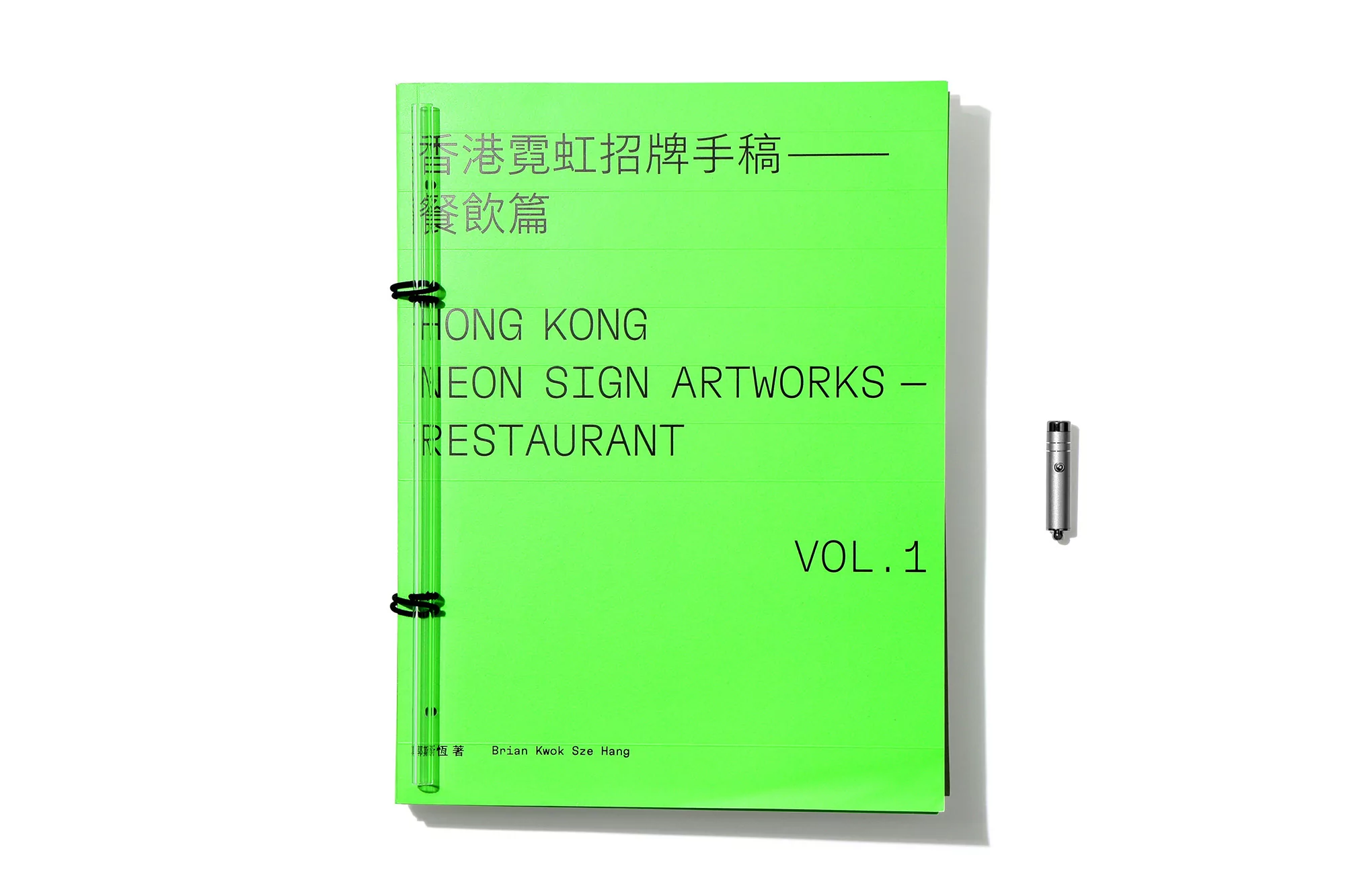 Bright green book with black spiral binding on a white surface next to a small silver metal object, featuring black text in English and Chinese.