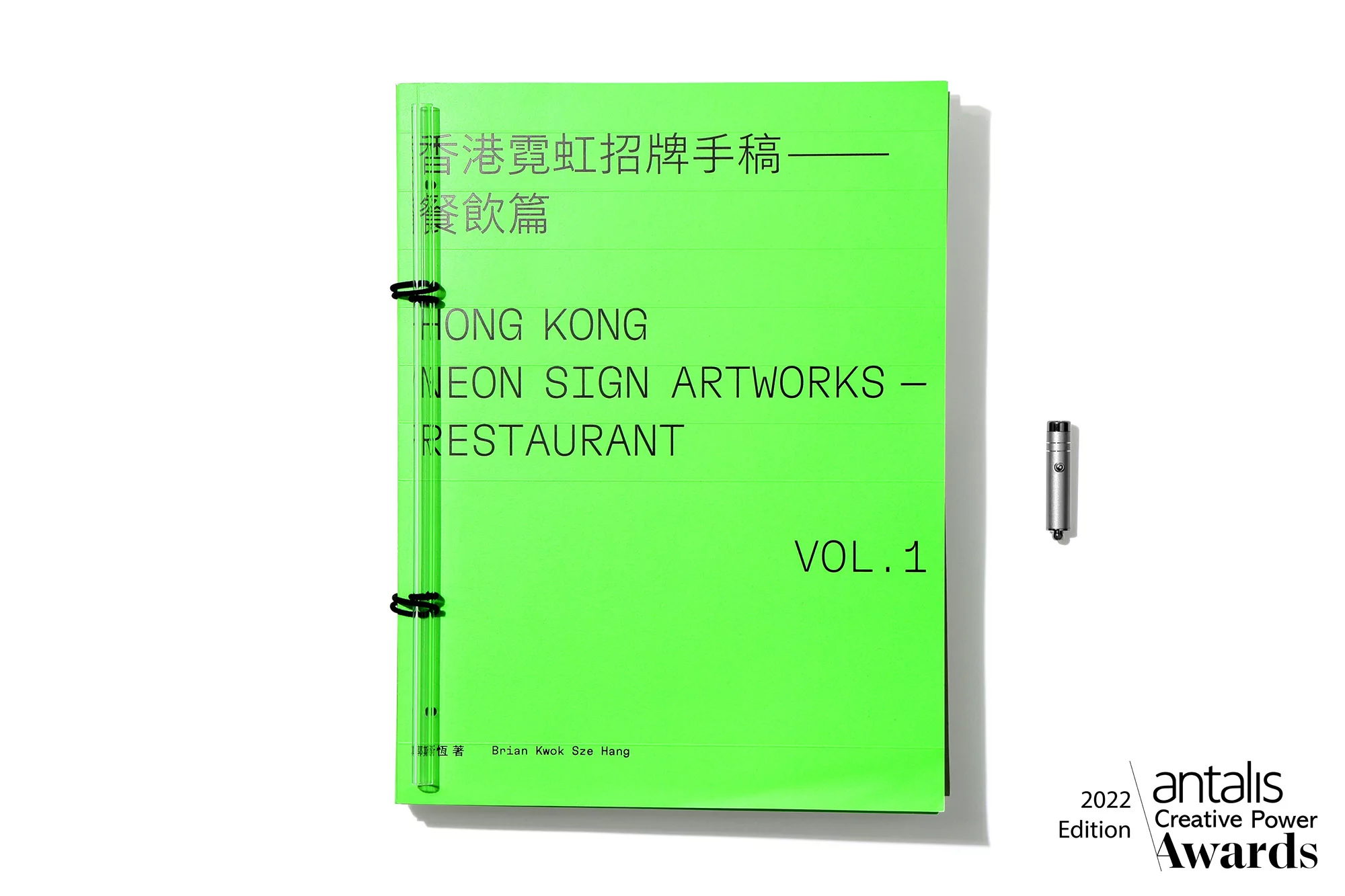 Bright green book titled 'Hong Kong Neon Sign Artworks - Restaurant Vol. 1' with black spiral binding, next to a small, silver metal object.