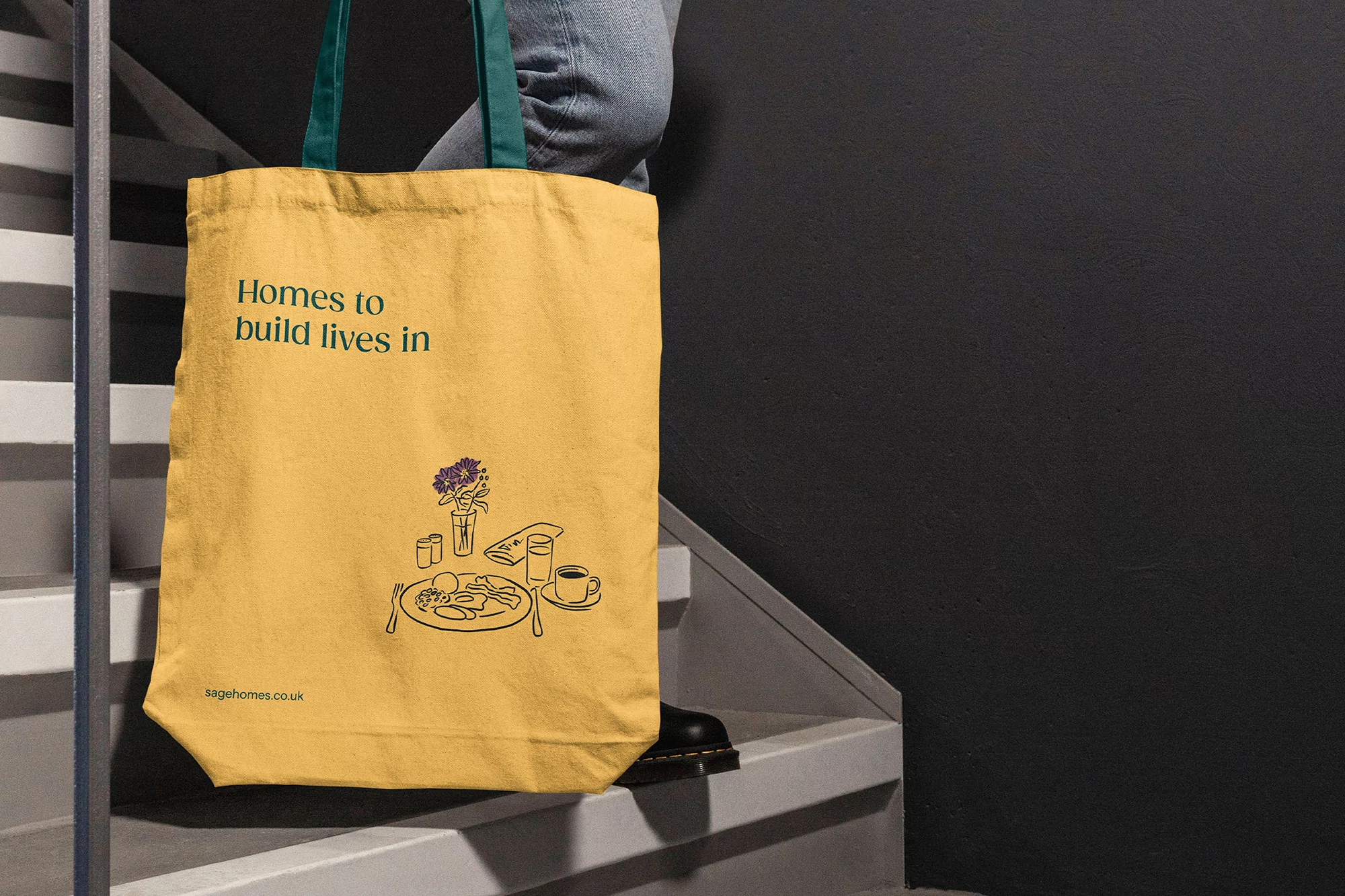 Tote bag on steps with 'Homes to build lives in' text and breakfast graphic design. Gold-colored bag with teal handle, steps and gray wall.
