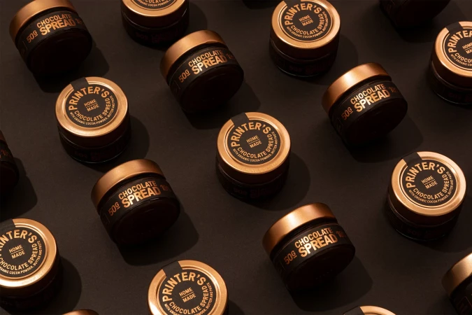 Overhead shot of multiple jars of Printer's chocolate spread with copper lids arranged on a brown surface in a grid-like pattern.