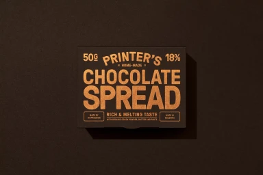 Top-down view of a rectangular black chocolate spread box with bold golden typographic branding on a dark textured paper background.