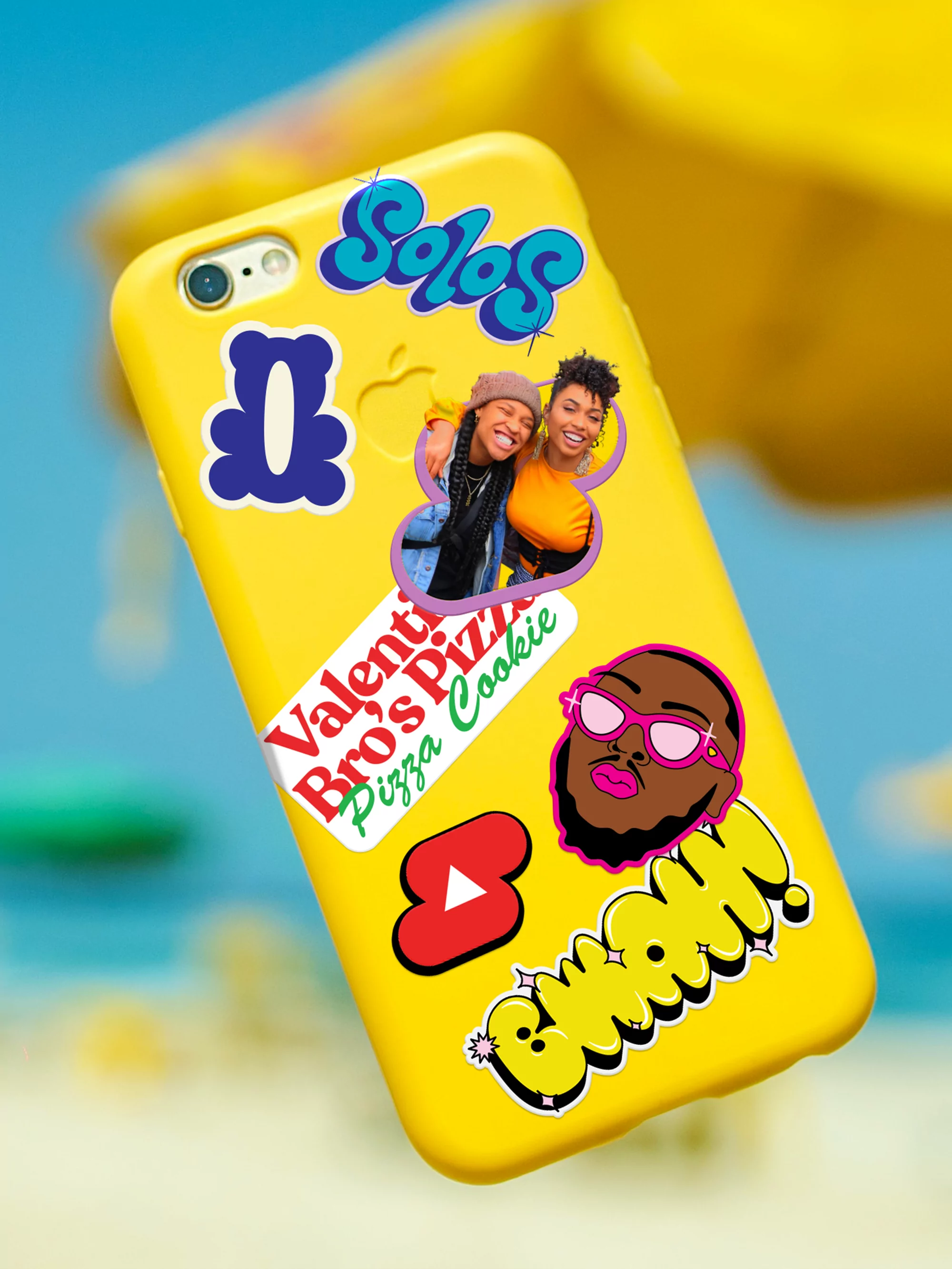 Yellow phone case covered in stickers, featuring two women in a photo and cartoon drawings against a blurred background.