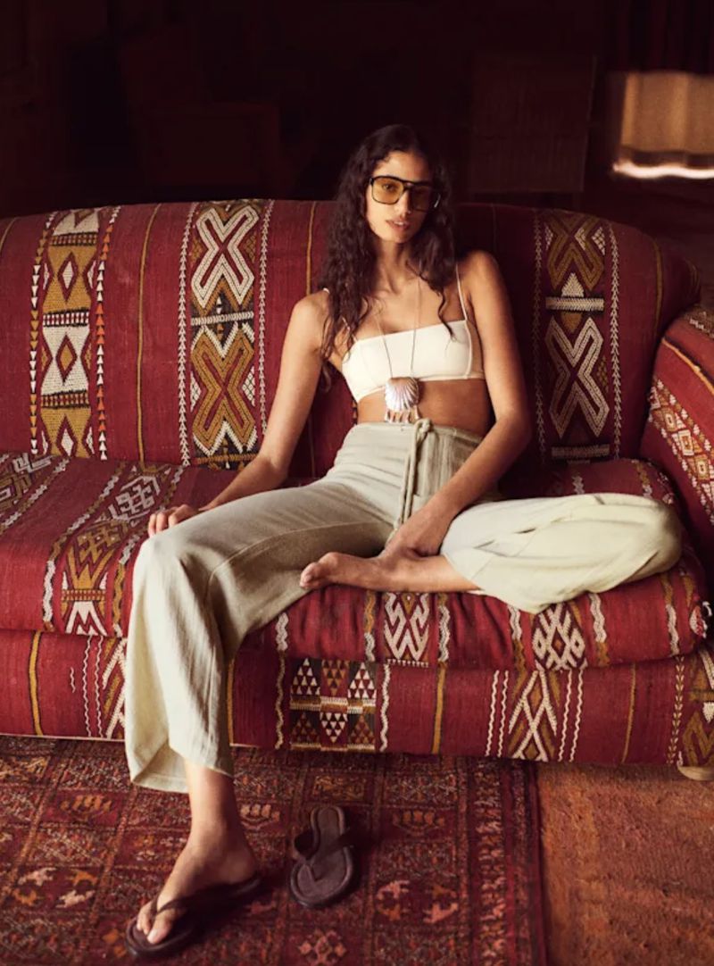 Adult female model sits on an ornate couch wearing a crop top, wide-leg pants, sunglasses, and sandals indoors with a patterned rug.