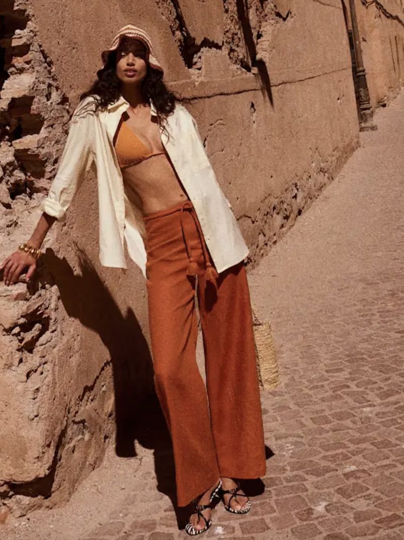 A woman in an orange bikini top and wide-leg pants leans against a textured wall outdoors, wearing a cream-colored shirt and bucket hat.