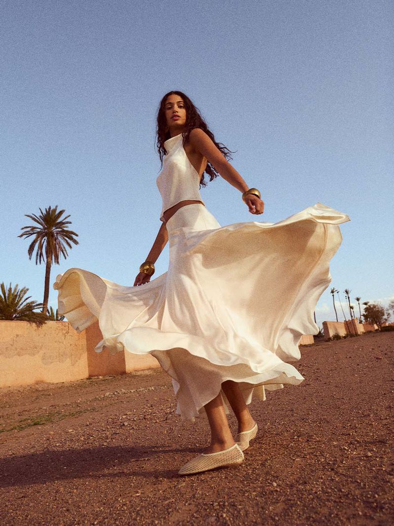 Woman in flowing white dress twirling outdoors in a desert-like setting with palm trees under a clear sky, captured in natural light.