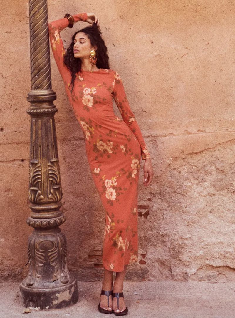 Woman in a floral print dress leans against a post on a peach-colored outdoor stone street.  She wears cuff bracelet and dangling earrings.