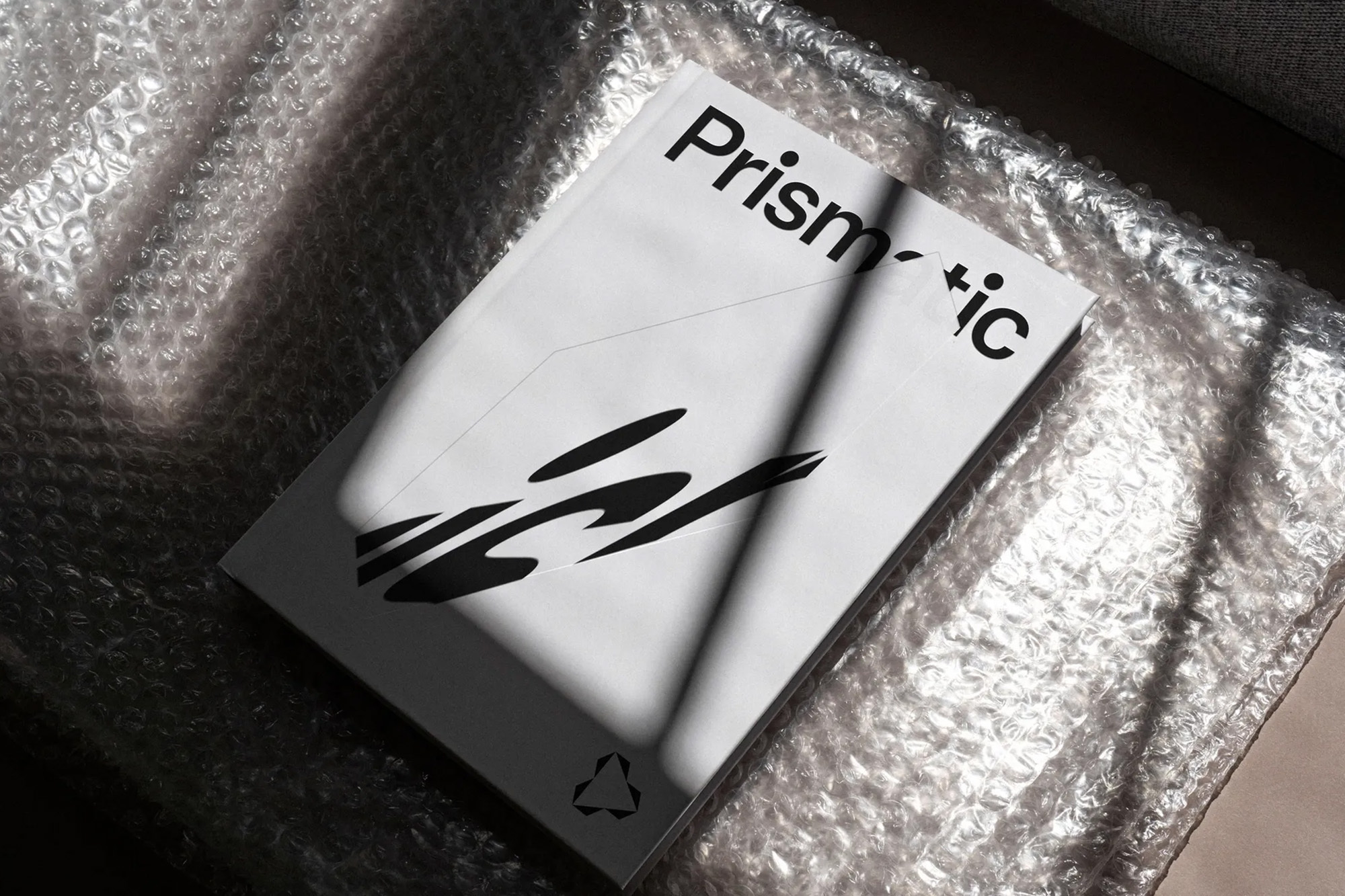 A modern, white hardcover book titled 'Prismatic' with black graphic elements, resting on translucent bubble wrap with natural sunlight.