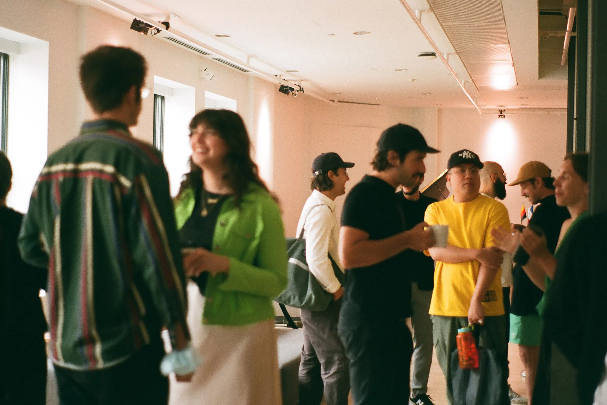 Indoor gathering of adults in casual wear, with conversations in a brightly lit room, featuring natural light from large windows.