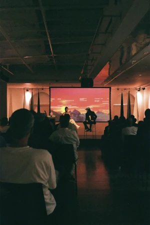 An audience seated in a darkened auditorium watching a presentation on a large projection screen displaying a landscape graphic