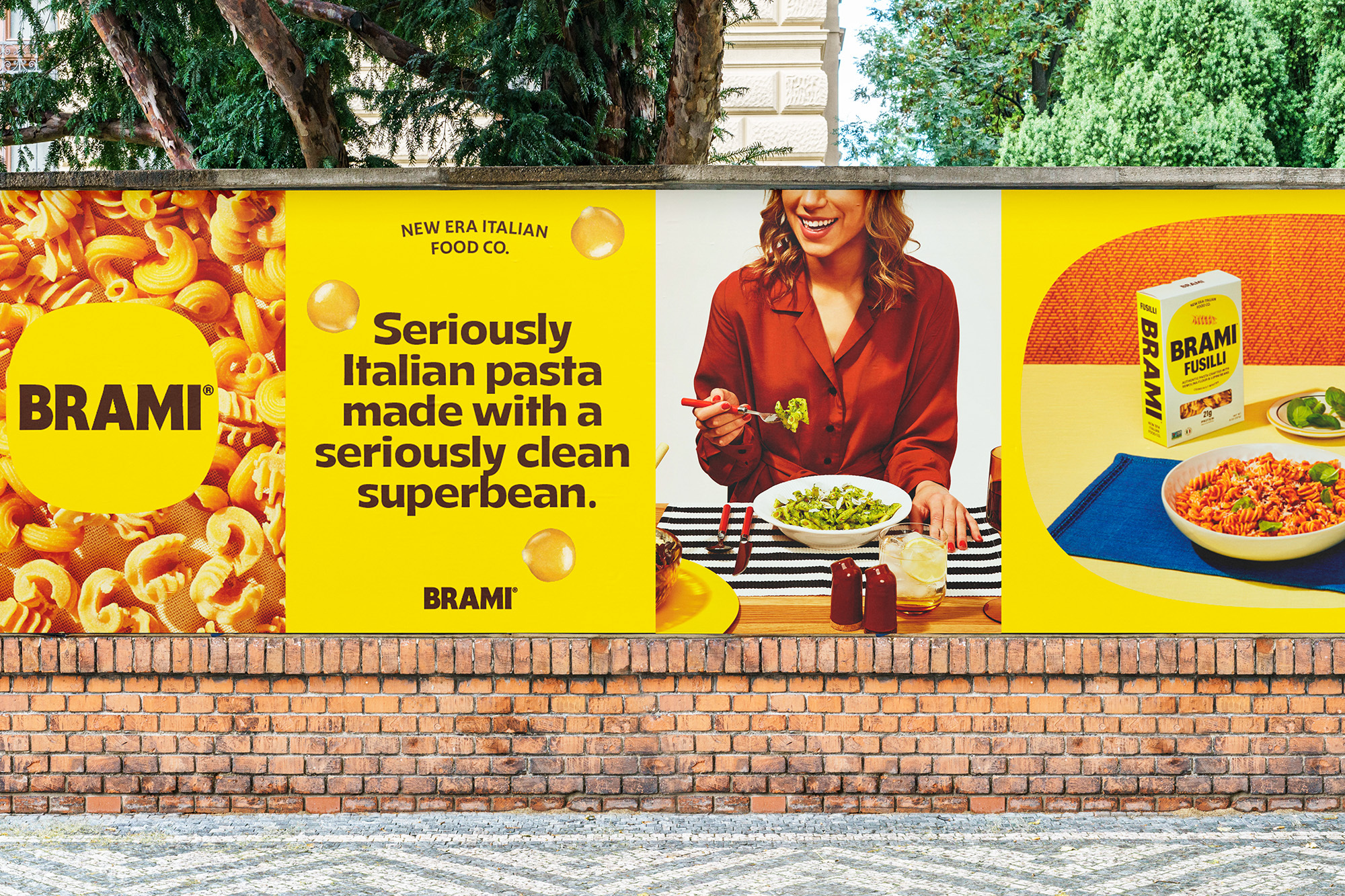 Eye-level shot of an outdoor advertisement for Brami pasta, featuring a woman eating pasta, and pasta dishes displayed on a brick wall.