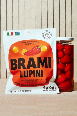 Brami lupini beans in a white and orange package, next to a glass jar of pickled peppers, against a striped backdrop.