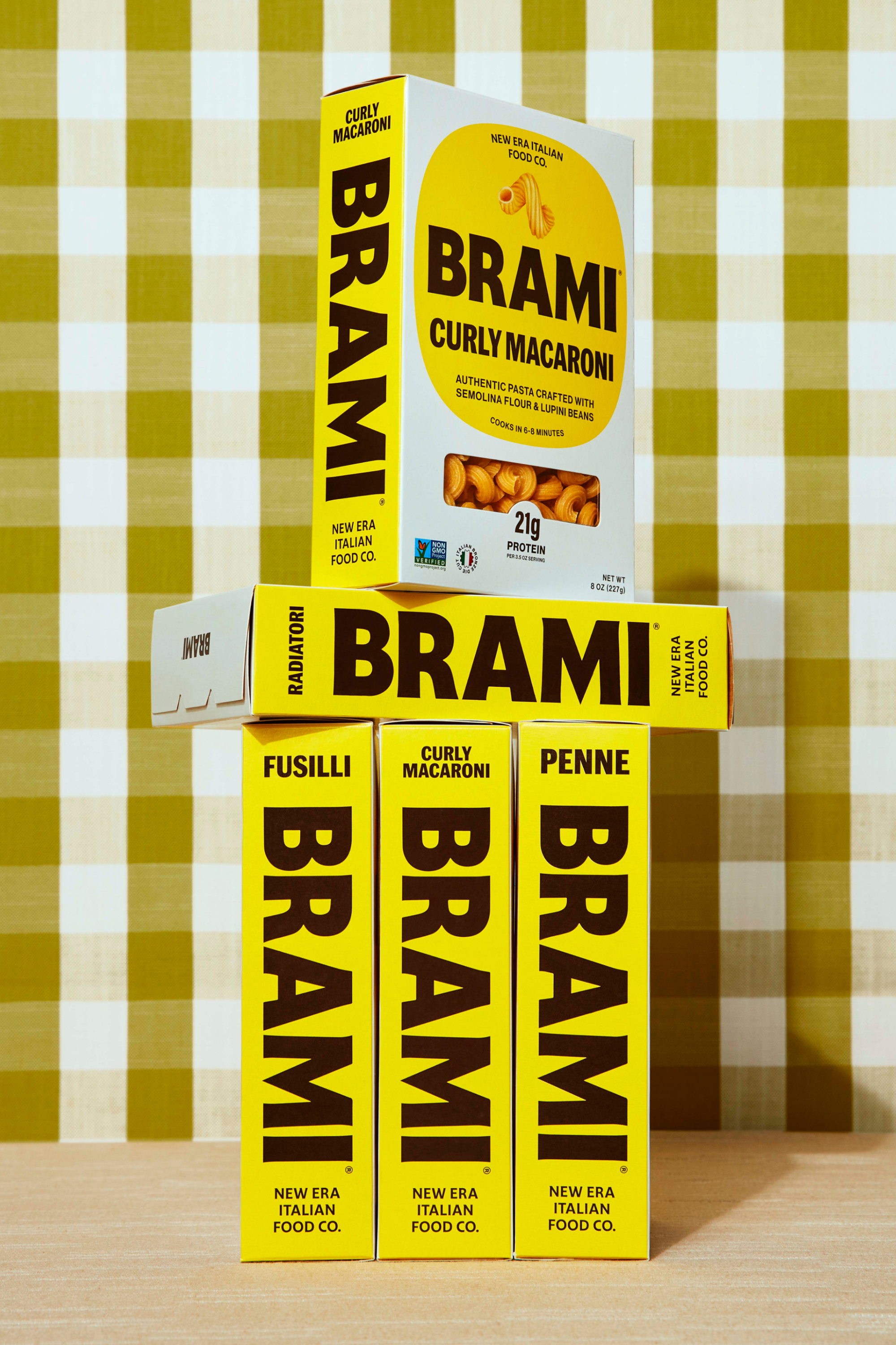 Several yellow boxes of Brami pasta sit on a surface against a checkered background, showing fusilli, penne, and curly macaroni.