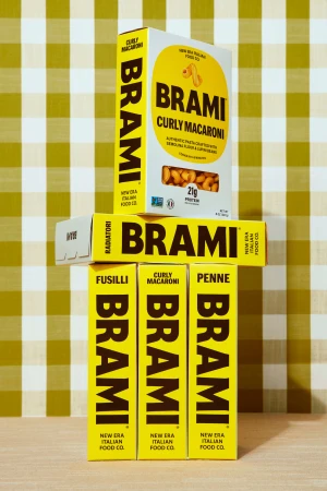 Several yellow boxes of Brami pasta sit on a surface against a checkered background, showing fusilli, penne, and curly macaroni.