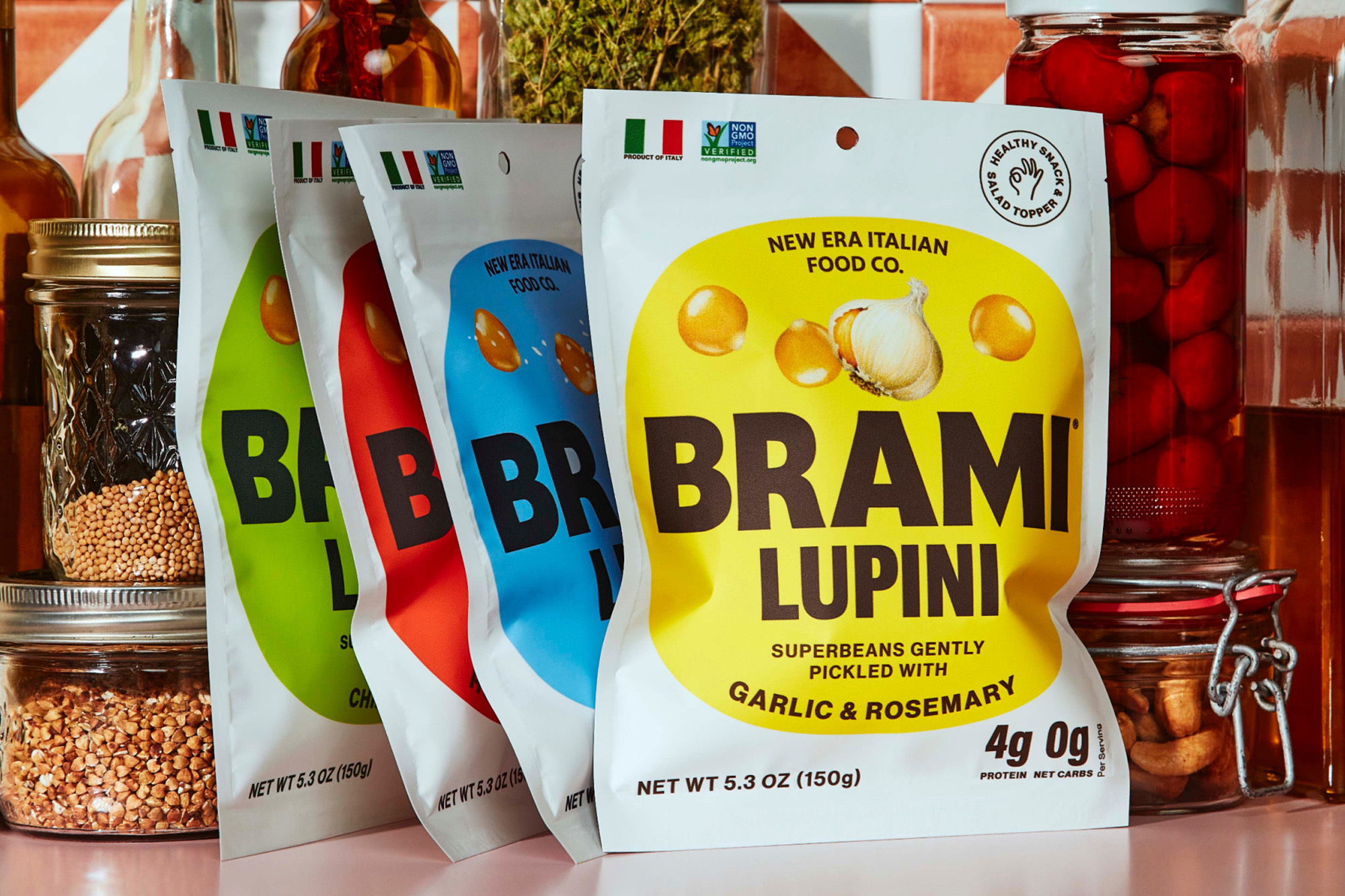 Four colorful packages of Brami Lupini beans arranged with glass jars and bottles against a tiled backsplash for food product photography.