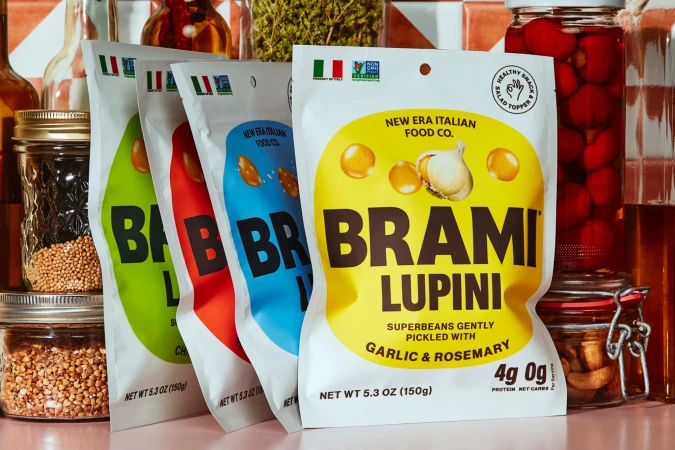 Four colorful packages of Brami Lupini beans arranged with glass jars and bottles against a tiled backsplash for food product photography.