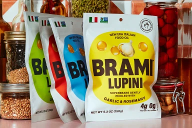 Four Brami Lupini snack pouches in green, red, blue, and yellow arranged in a row with glass jars of preserved ingredients in the background.