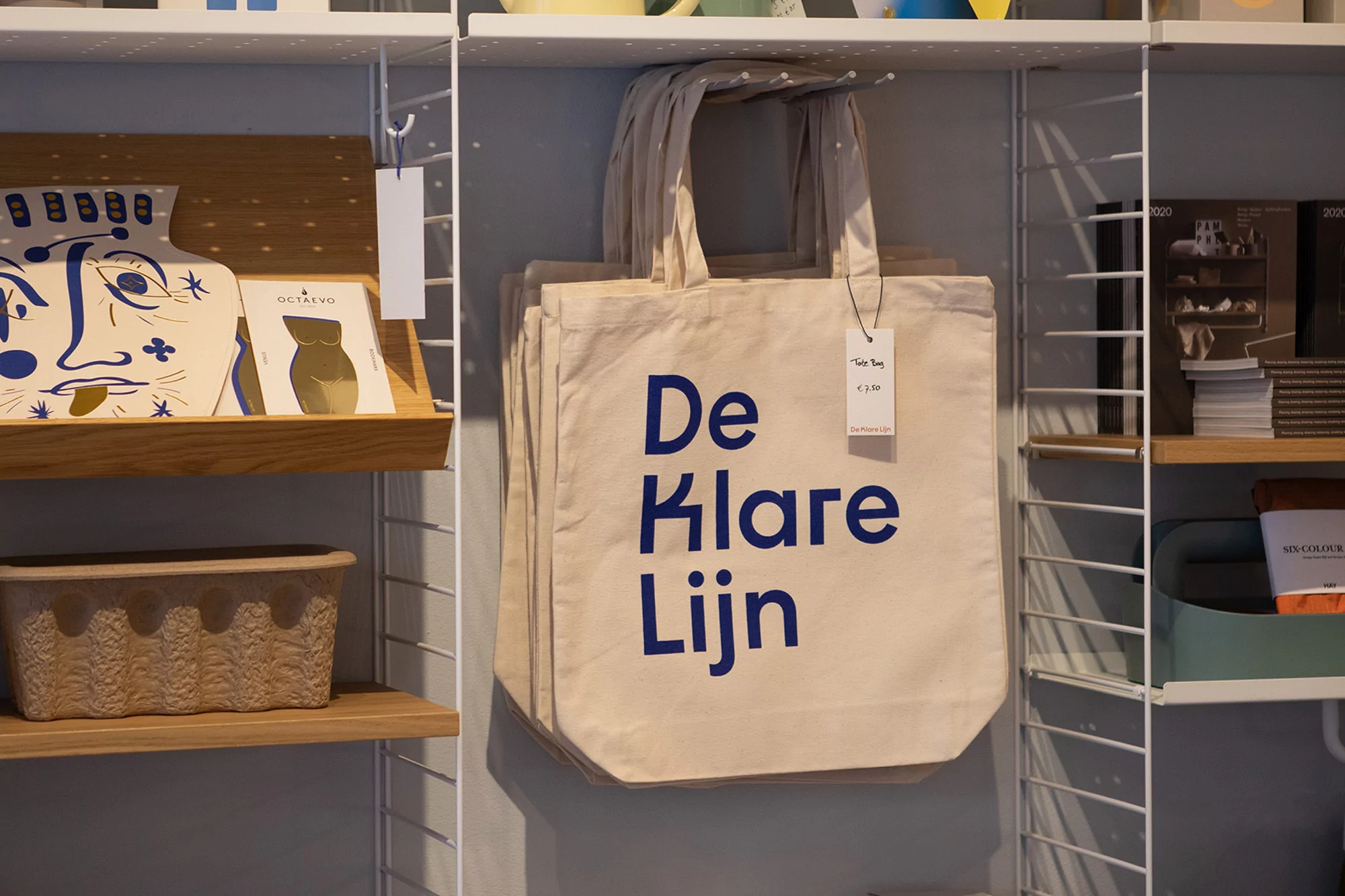 Display of beige canvas tote bags with blue text, stacked on a shelf, alongside various items in a retail setting.